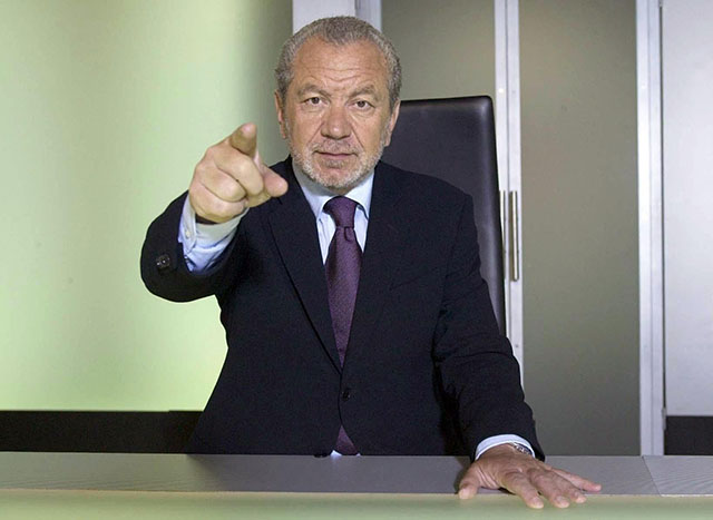 Dressed in a black suit and sitting at a desk, The Apprentice's Lord Alan Sugar