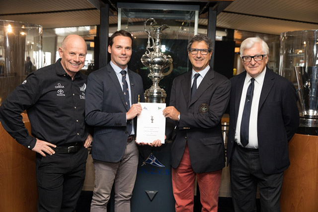 Four men hold a piece of paper in front of the silver America's Cup